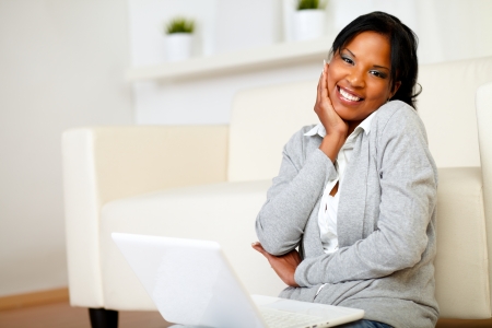 Portrait of a cute black woman smiling at you while is sitting on the floor with laptopの写真素材