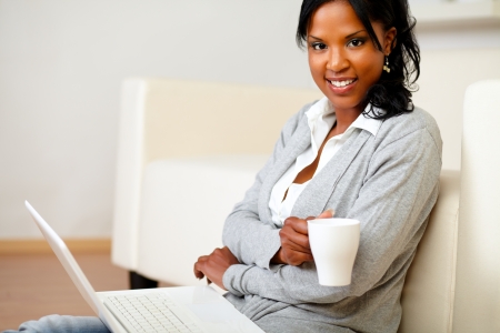 Portrait of a young woman with a mug is looking to you and browsing the Internet on her laptop while is sitting on the floor at home indoorの写真素材