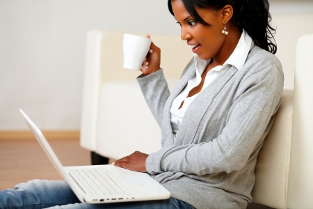 Portrait of a pretty woman with a mug browsing the Internet on her laptop while is sitting on floor at home indoorの写真素材