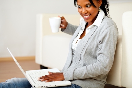 Portrait of a young woman with a mug browsing the Internet on her laptop while is sitting on the floor at home indoorの写真素材