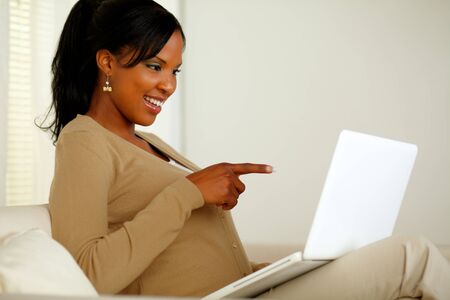 Portrait of a charming woman pointing and looking to laptop screen while sitting on sofaの写真素材