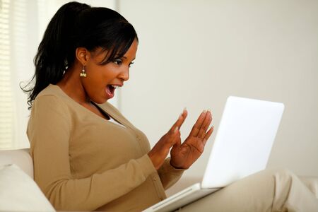 Portrait of an excited woman looking to laptop screen while sitting on sofa at home indoorの写真素材