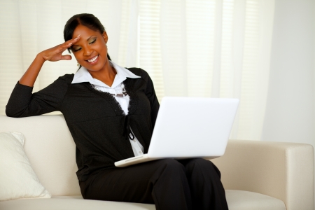 Portrait of a charming executive black woman reading on laptop screen. Sitting on a couch and laughing and having fun while working at homeの写真素材