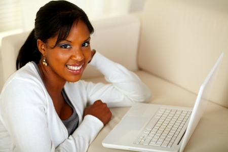 Portrait of a charming young woman working on laptop at home indoor while looking and smiling at youの写真素材