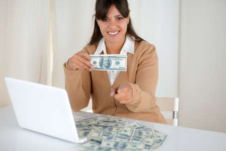 Portrait of a young woman holding and showing you cash dollars in front of her laptop while is...