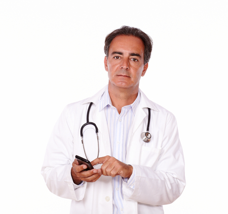 A portrait of an adult charismatic doctor on white uniform sending a message using his mobile while looking at you on isolated backgroundの写真素材