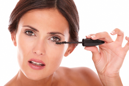 Front view portrait of pretty woman using make up brush to her eyelash with nude shoulders while looking at camera on isolated studioの写真素材