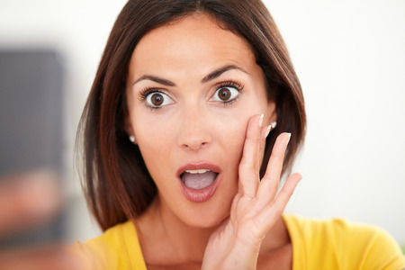 Close up portrait of a young woman looking surprised while using a mobile phone against white backroundの写真素材