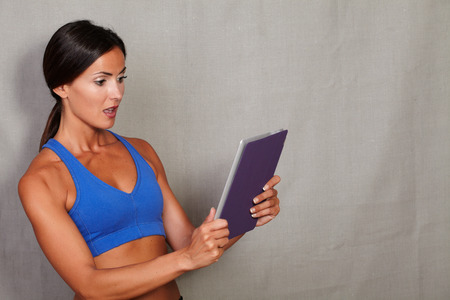 Surprised woman holding tablet with open mouth and wearing fitness clothing on grey texture backgroundの写真素材