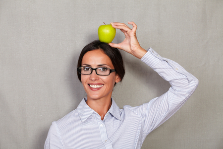Young businesswoman with glasses holding apple on head while smiling and looking at camera in button down shirt against grey texture backgroundの写真素材