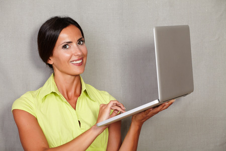 Long hair female in green blouse holding laptop and smiling with toothy smile while looking at camera on grey texture backgroundの写真素材