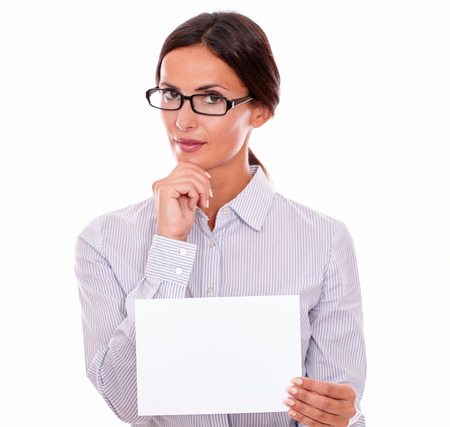 Indifferent brunette businesswoman with glasses, wearing her long hair tied back, and a button down shirt, holding a blank signboard in one hand, and the other hand on her chinの写真素材
