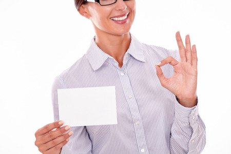 Smiling brunette businesswoman with glasses, wearing her long hair tied back, and a button down shirt, holding a blank copy space in one hand gesturing a perfect sign with the other handの写真素材