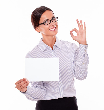 Satisfied brunette businesswoman with glasses, wearing her long hair tied back, and a button down shirt, holding a blank copy space in one hand, gesturing perfect with the other handの写真素材