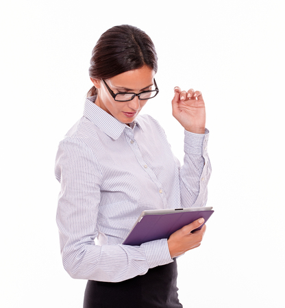 Interested, concentrating brunette businesswoman with glasses looking at a tablet, wearing her straight hair tied back and a button down shirt, with her left hand up, on a white backgroundの写真素材