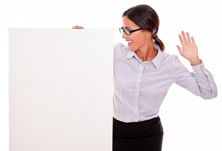 Surprised brunette businesswoman with glasses looking at a placard with her open mouth, wearing her straight hair tied back and a button down shirt, gesturing with her left hand in the airの写真素材