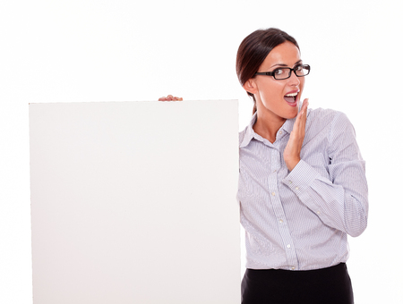 Impressed brunette businesswoman looking at the camera, holding a placard with her open mouth, wearing her straight hair tied back and a button down shirt, with a gesture of her left hand to her mouthの写真素材