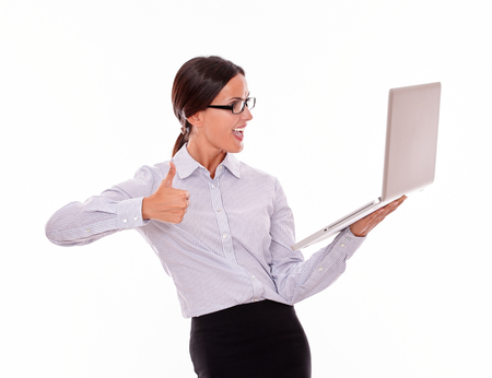 Smiling brunette businesswoman looking at laptop very happy while making a thumb up gesture with one hand and wearing her straight hair back in a button down shirt from waist up on a white backgroundの写真素材