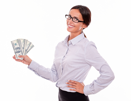 Happy smiling brunette businesswoman holding money with one hand on her hip while looking at the camera and wearing her straight hair back in a button down shirt on a white backgroundの写真素材