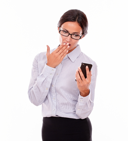 Shocked businesswoman looking at her cell phone making a surprised gesture with one hand to her mouth while wearing her straight hair back and a button down shirt on a white backgroundの写真素材