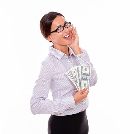 Excited brunette businesswoman holding money with an impressed gesture of her hand to her face while wearing her straight hair back and a button down shirt from the waist up on a white backgroundの写真素材