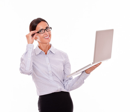 Smiling brunette businesswoman holding a laptop with a toothy smile while looking at the camera and wearing her straight hair back while she is touching her glasses with one hand, isolatedの写真素材