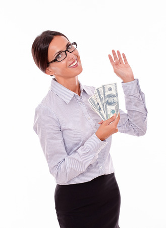 Satisfied and smiling brunette businesswoman looking at camera while holding money with a happy gesture and wearing her straight hair back in a button down shirt from waist up on a white backgroundの写真素材