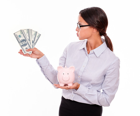 Satisfied businesswoman holding pink porcelain piggy bank in one hand and money in the other hand while blowing a kiss at the money and wearing her hair back on a white backgroundの写真素材