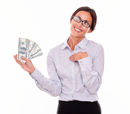 Happy smiling brunette businesswoman holding dollar bills while smiling and looking away, marveling with one hand on her chest in a button down shirt and her hair tied back on a white backgroundの写真素材