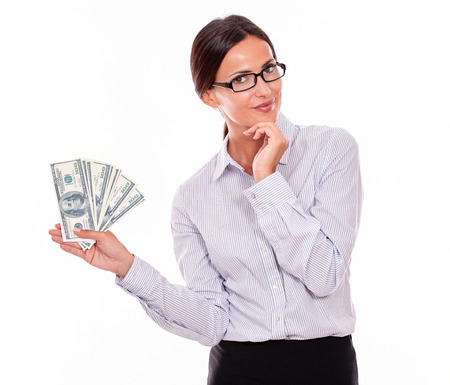 Thinking smiling brunette businesswoman holding dollar bills while smiling and looking at the camera, marveling with one hand on her chin in a button down shirt and her hair tied back, isolatedの写真素材