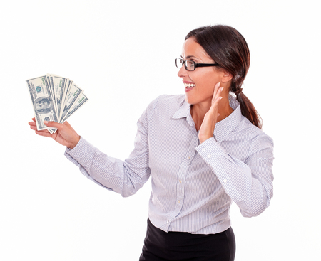 Impressed brunette businesswoman holding dollar bills with an excited gesture of one hand and smiling while looking at money in a button down shirt and her hair tied back on a white backgroundの写真素材