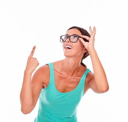 Surprised brunette woman with green tank top looking up and pointing upwards to copy space with one hand while smiling and touching her glasses with her long hair back on a white backgroundの写真素材
