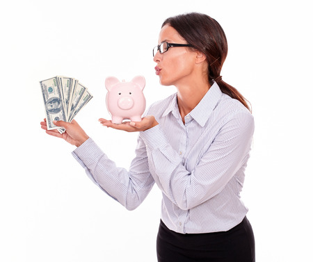 Businesswoman holding pink porcelain piggy bank in one hand and money with the other hand while blowing a kiss at the piggy bank and wearing her hair back in a button down shirt on a white backgroundの写真素材
