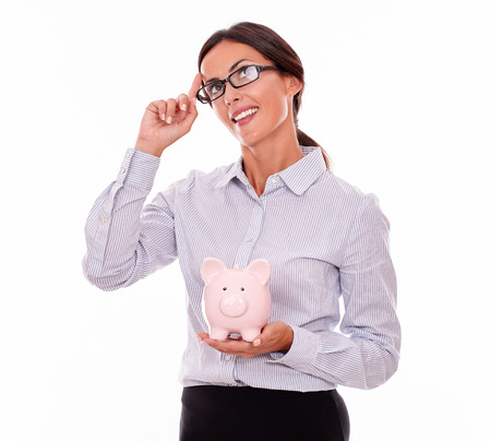 Thinking businesswoman holding pink porcelain piggy bank smiling and looking at the sky while wearing her hair back and a button down shirt with a planning gesture of one hand on a white backgroundの写真素材