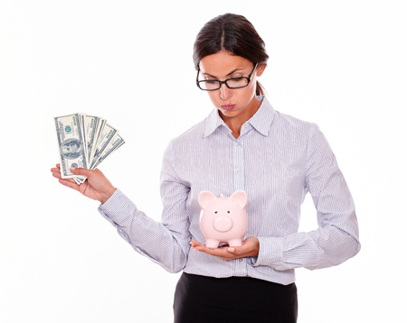 Businesswoman holding pink porcelain piggy bank in one hand and money with the other hand while looking satisfied at the piggy bank wearing her hair back in a button down shirt on a white backgroundの写真素材