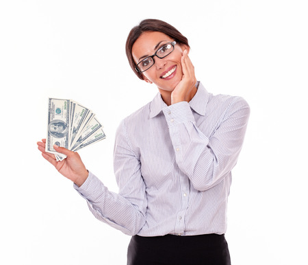 Happy smiling brunette businesswoman holding dollar bills while smilingly looking at the camera and marveling with one hand on her chin wearing a button down shirt and her hair tied back, isolatedの写真素材