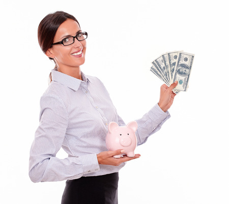 Businesswoman holding pink porcelain piggy bank in one hand and money with the other hand showing saving money while wearing her hair back and smiling at camera on a white backgroundの写真素材