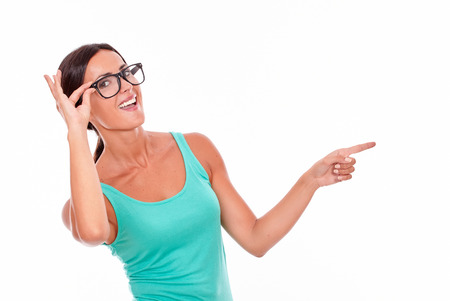 Pointing brunette woman in green tank top looking at camera with a toothy smile while pointing at copy space with one hand and smiling while touching her glasses and her long hair back, isolatedの写真素材