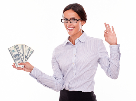 Happy brunette businesswoman holding dollar bills making a perfect gesture and smilingly looking at the camera while wearing a button down shirt and her hair tied back on a white backgroundの写真素材