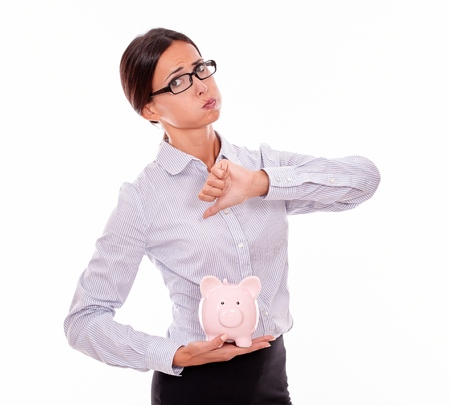 Businesswoman holding pink porcelain piggy bank with disapproval and a thumb down gesture wearing her hair back and a button down shirt while looking at camera on a white backgroundの写真素材