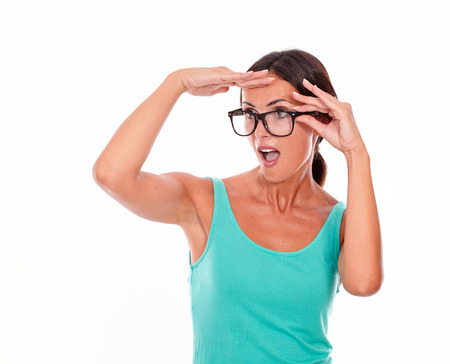 Shocked adult woman with a hand to her forehead touching her glasses with the other hand while looking into the distance wearing her long hair tied back in a green tank top on a white backgroundの写真素材