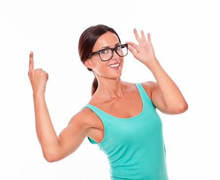 Adult woman pointing at copy space above her head while looking at the camera with a confident smile and wearing her long hair tied back in a green tank top touching her glasses on a white backgroundの写真素材