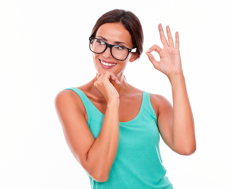 Satisfied smiling adult woman looking away while showing an ok sign and touching her chin wearing her long hair tied back in a green tank top on a white backgroundの写真素材