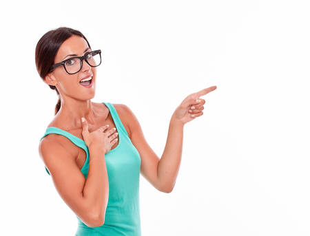 Surprised woman pointing at copy space next to her while looking at the camera with one hand on her chest and wearing her long hair tied back in a green tank top on a white backgroundの写真素材
