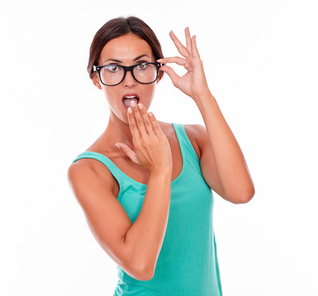 Surprised adult woman with a hand to her mouth while showing touching her glasses with the other wearing her long hair tied back in a green tank top on a white backgroundの写真素材