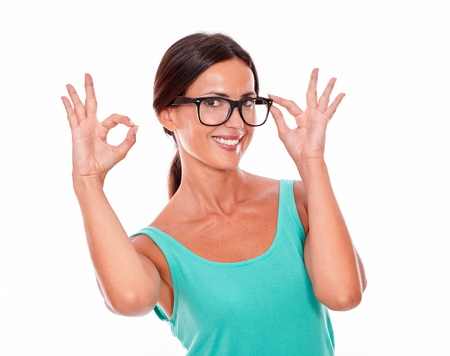 Satisfied adult woman smiling at camera while showing an ok sign with one hand and touching her glasses with the other wearing her long hair tied back in a green tank top on a white backgroundの写真素材
