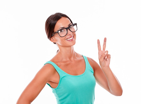 Celebrating adult woman smiling at camera while showing a victory sign with one hand and wearing her long hair tied back in a green tank top on a white backgroundの写真素材