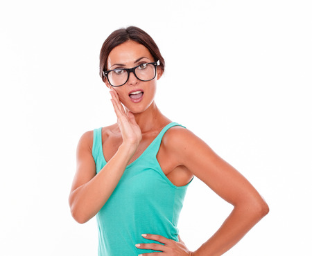 Surprised adult woman holding her chin while looking at the camera with an open mouth and wearing her long hair tied back in a green tank top on a white backgroundの写真素材