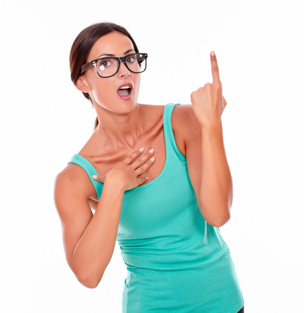 Surprised woman pointing at copy space above her head while looking at the camera with one hand on her chest and wearing her long hair tied back in a green tank top on a white backgroundの写真素材