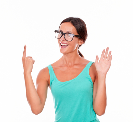 Adult woman pointing at copy space above her head while looking at the camera with a charismatic smile and wearing her long hair tied back in a green tank top on a white backgroundの写真素材
